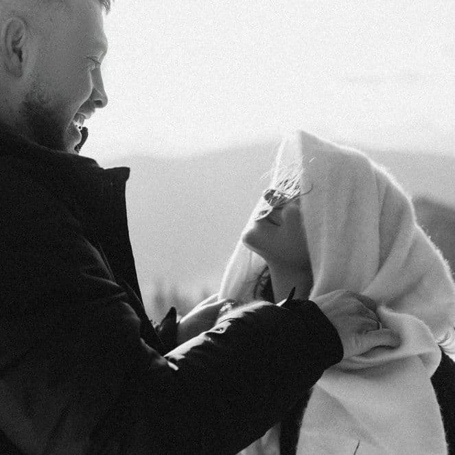 A man and woman smiling at each other on a snowy mountain trail. The woman is wrapped in a blanket. Black and white photo.