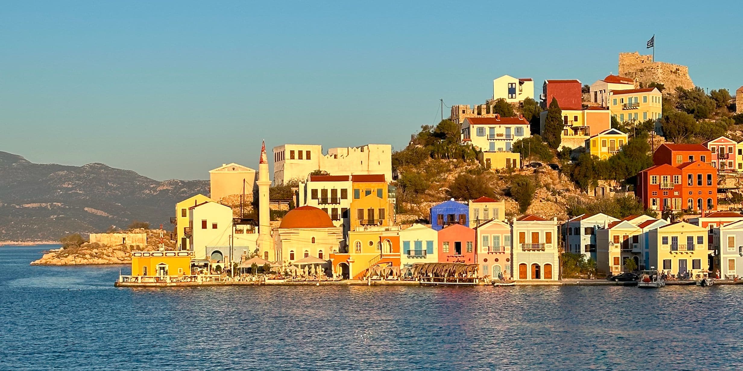 Colorful houses on a sunlit hillside by the sea, with a clear blue sky and distant mountains in the background.