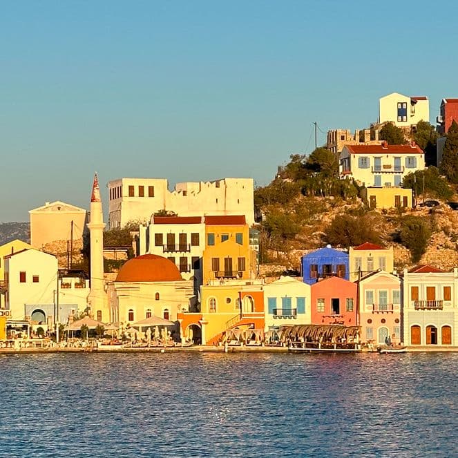 Colorful houses on a sunlit hillside by the sea, with a clear blue sky and distant mountains in the background.