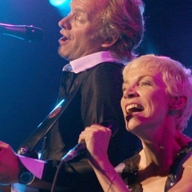 Two performers singing passionately on stage, one playing guitar. Blue stage lights illuminate the scene.