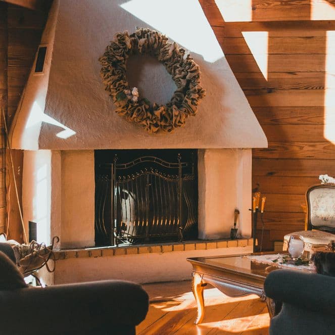 Cozy living room with a white fireplace, wreath, wooden walls, and sunlight streaming through large windows onto sofas and a coffee table.