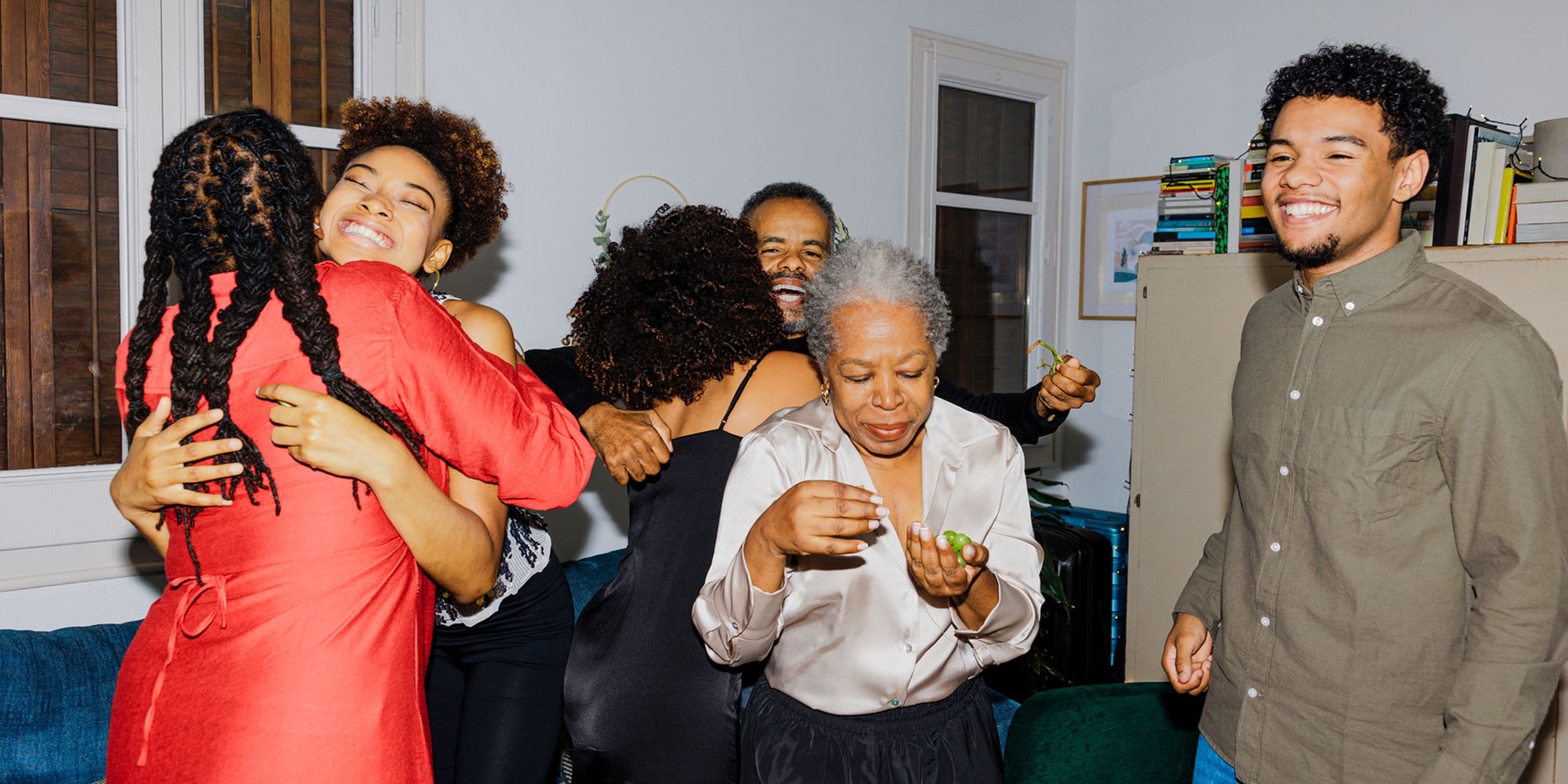 A joyful family gathering with people hugging and smiling in a small room, featuring a woman in a red dress and books on a shelf.