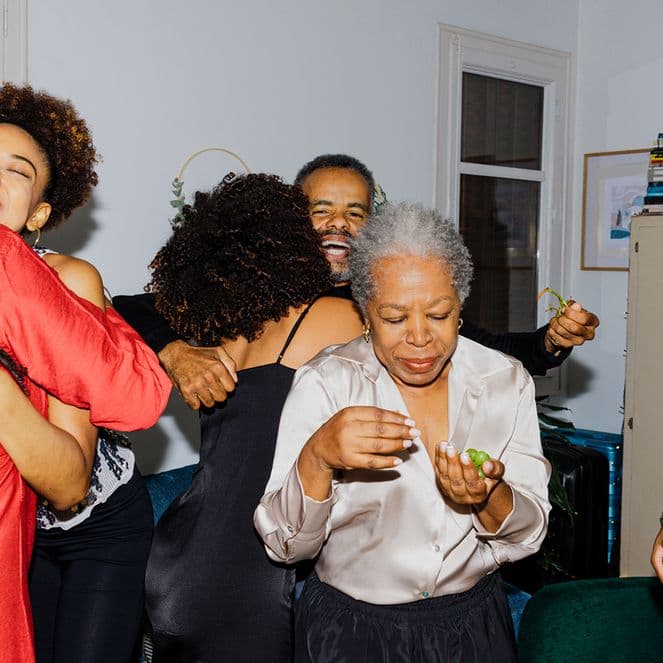 A joyful family gathering with people hugging and smiling in a small room, featuring a woman in a red dress and books on a shelf.