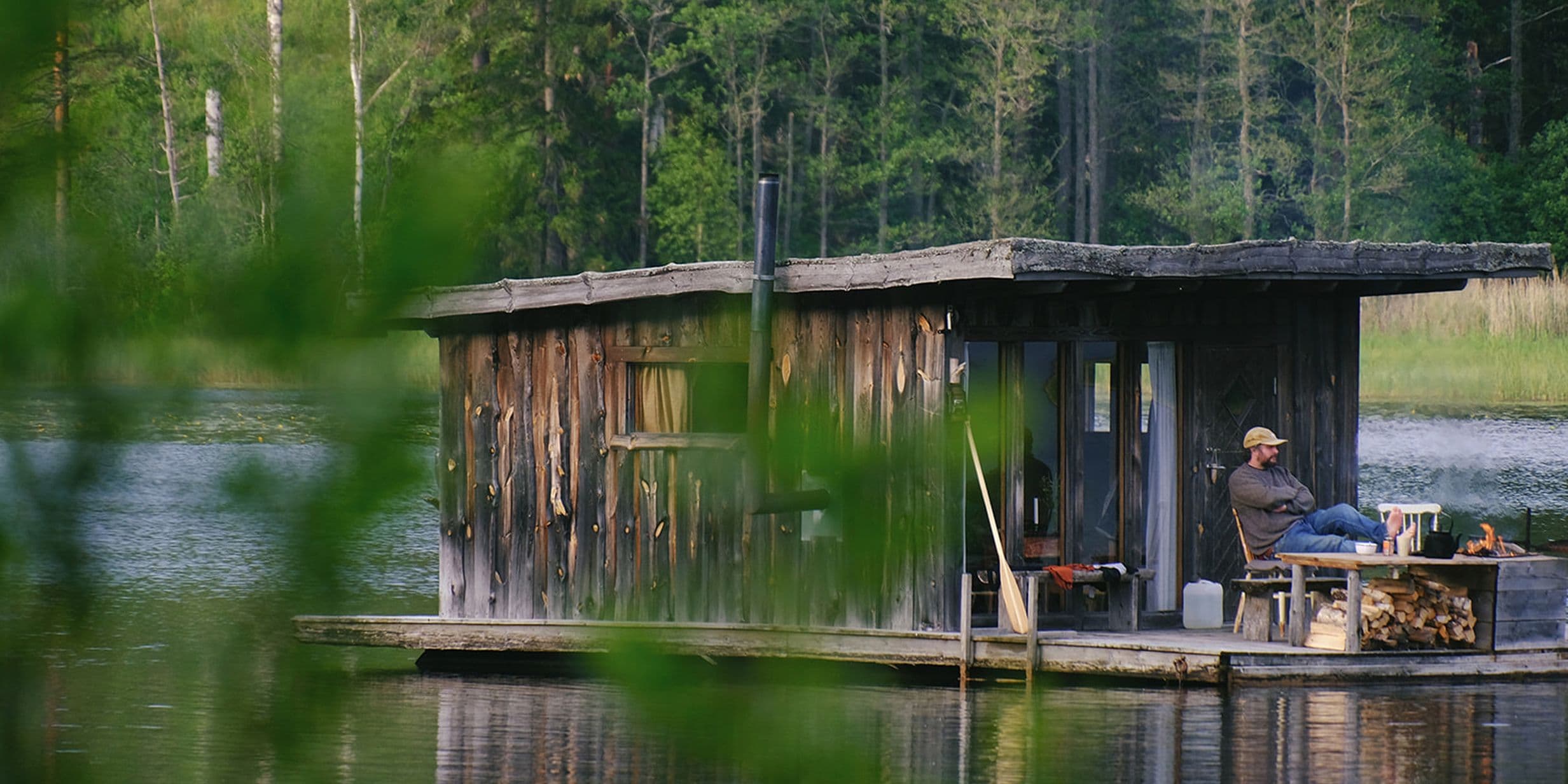 Man sitting on the deck of a rustic floating wooden cabin on a calm forest lake, blurred leaves in the foreground.