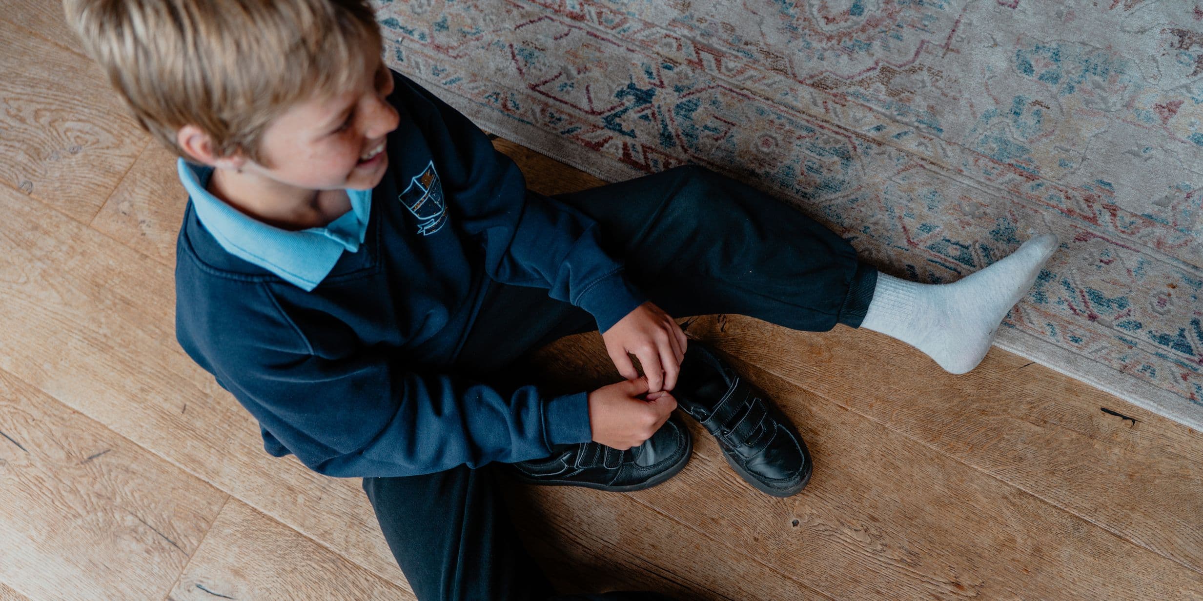 A child sits on a wooden floor, wearing a school uniform, tying the laces of one shoe while the other foot is covered in a white sock.