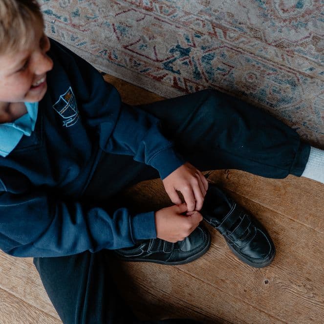 A child sits on a wooden floor, wearing a school uniform, tying the laces of one shoe while the other foot is covered in a white sock.