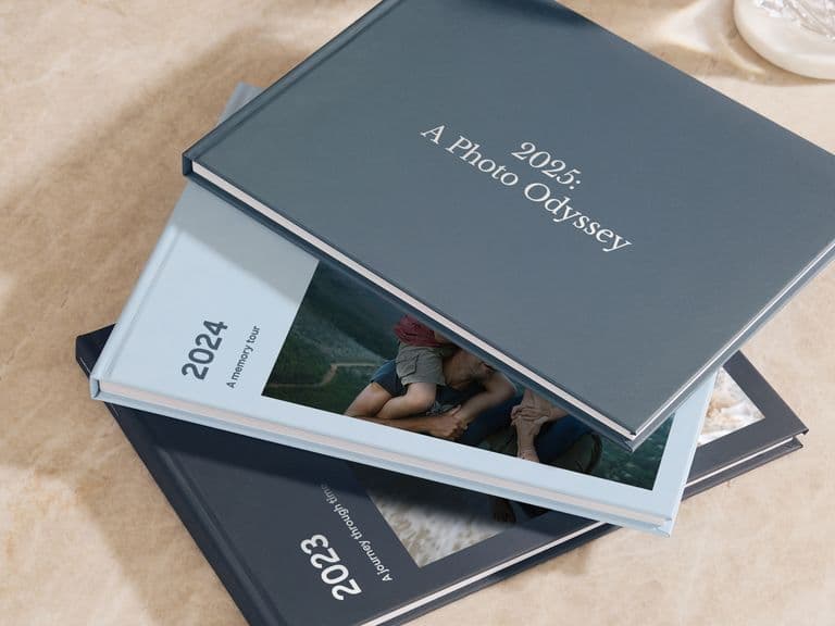 Three photo books titled "2023," "2024," and "2025: A Photo Odyssey" are stacked on a marble surface.