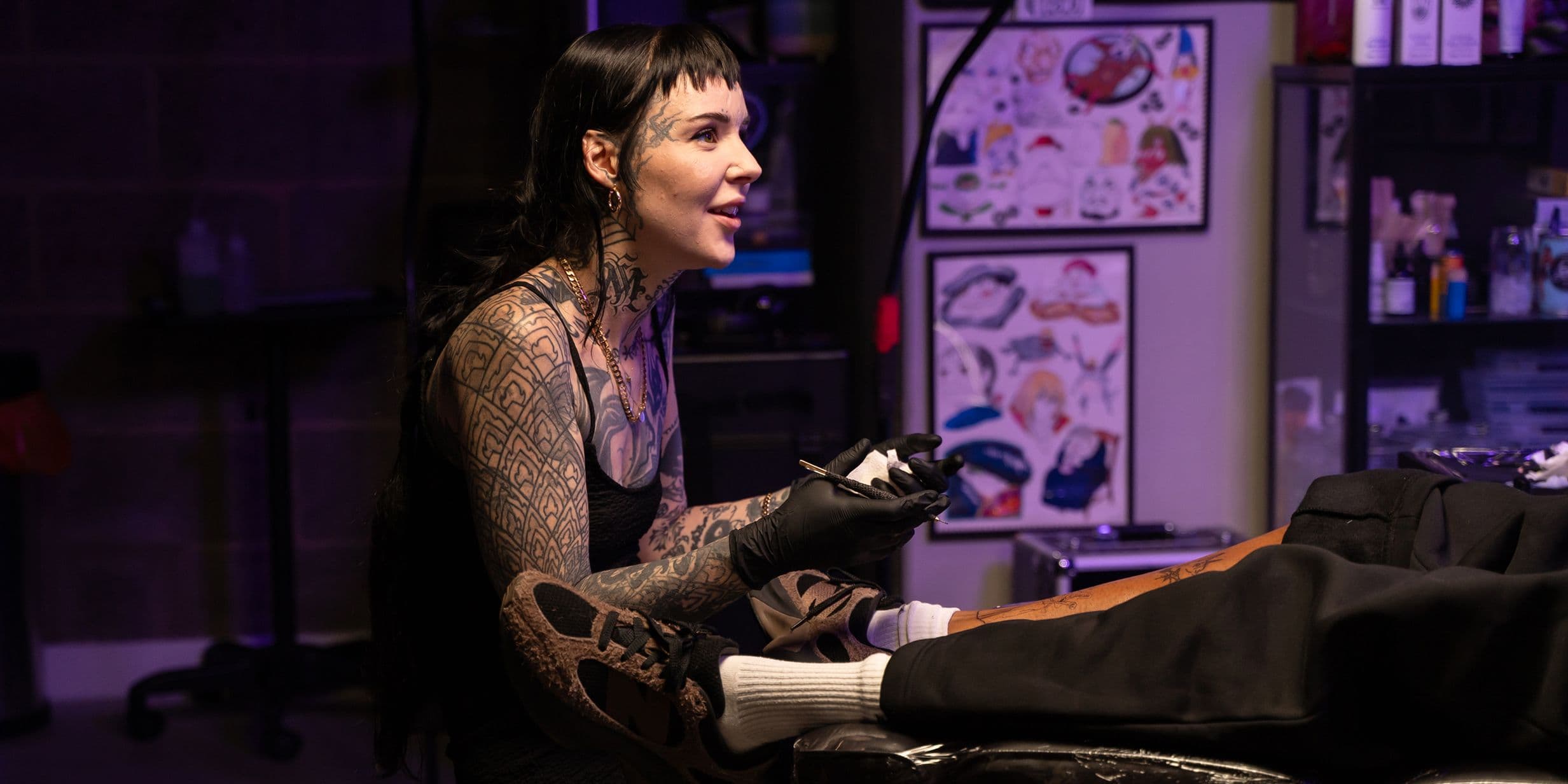 A tattoo artist with long dark hair and tattoos on her arms and face works on a client's leg in a dimly lit studio.