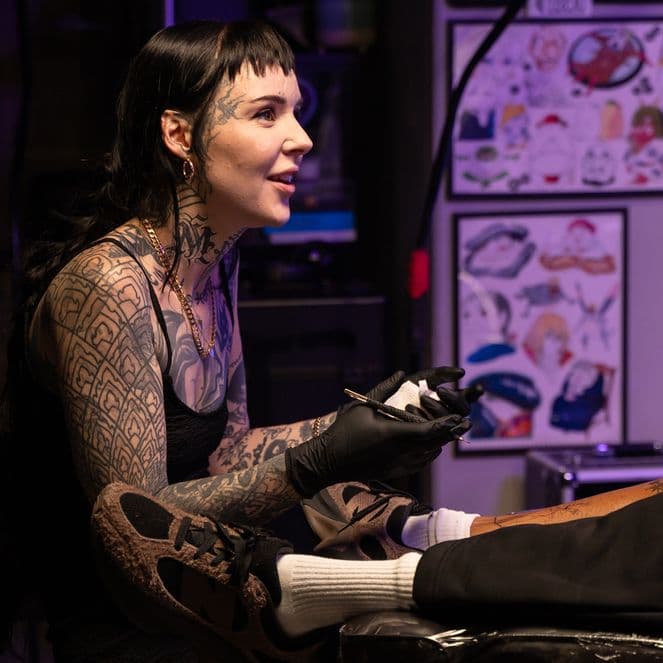 A tattoo artist with long dark hair and tattoos on her arms and face works on a client's leg in a dimly lit studio.