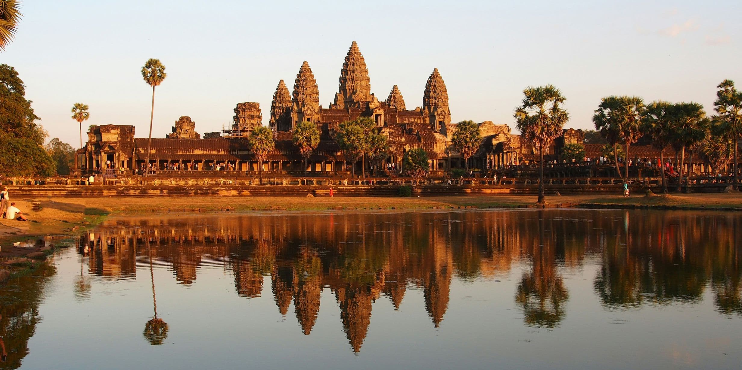 Angkor Wat temple complex at sunset, reflected in a calm pond, surrounded by palm trees and a clear sky.
