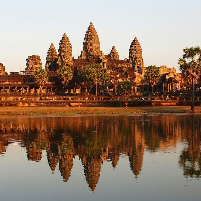 Angkor Wat temple complex at sunset, reflected in a calm pond, surrounded by palm trees and a clear sky.