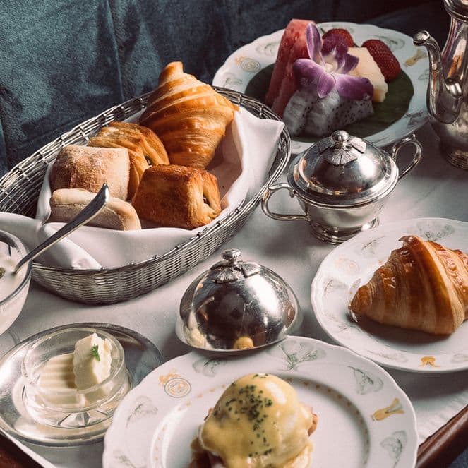 A breakfast tray with croissants, rolls, yogurt, butter, a poached egg dish, fruit salad, and a cup of coffee, set on a plush surface.