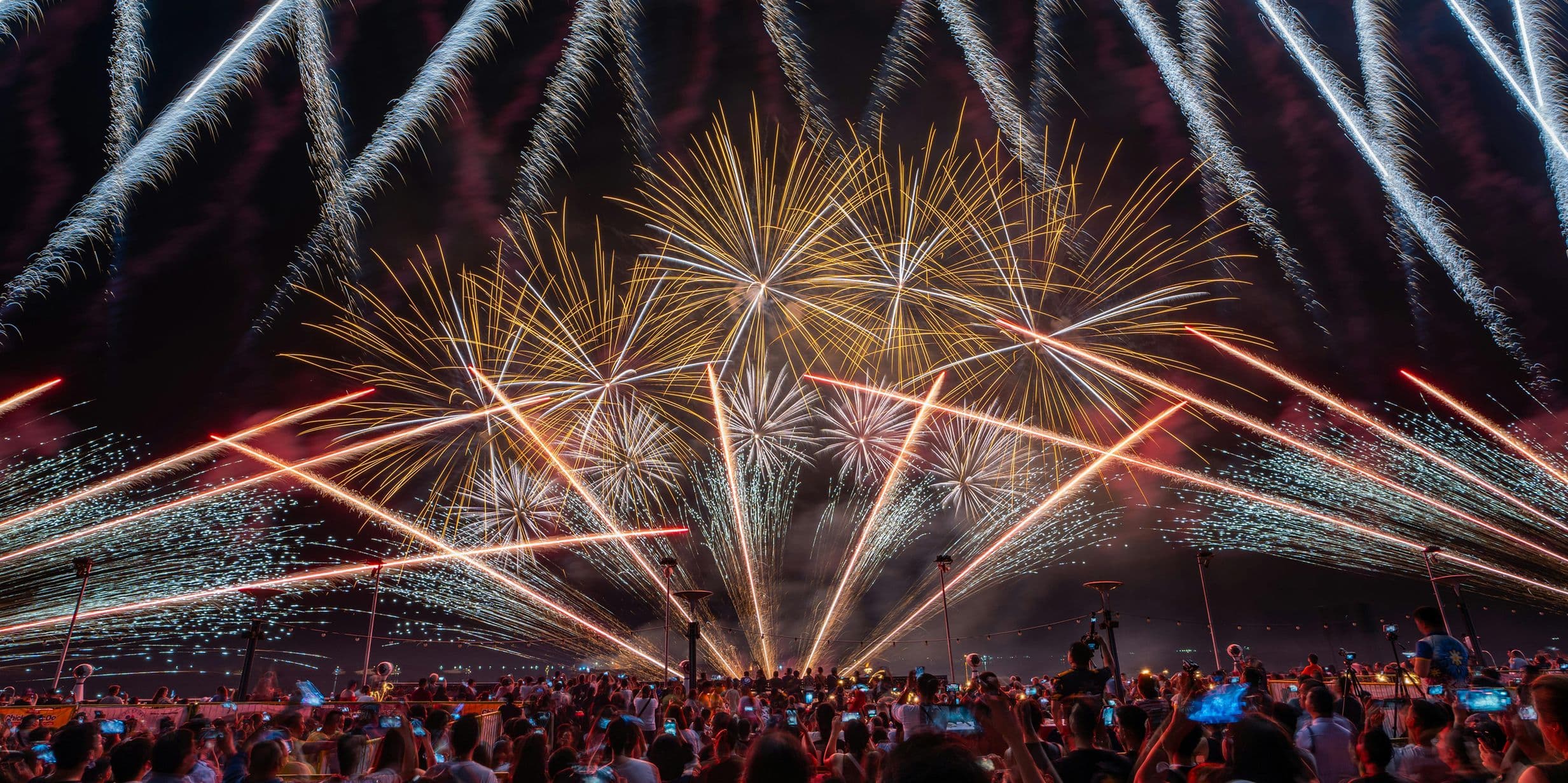 A vibrant fireworks display lights up the night sky, with a crowd of people watching and capturing the moment on their phones.