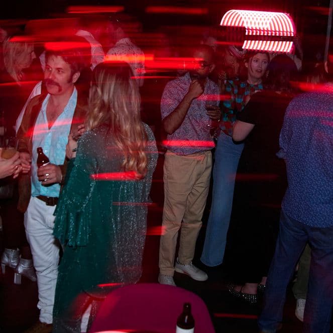 People at a lively party with red light streaks, wearing casual and semi-formal attire, some holding drinks, chatting, and mingling.