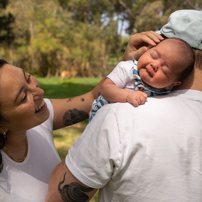 A woman smiles at a baby resting on a man's shoulder outdoors in a park. The baby is wearing a patterned outfit and a cap.