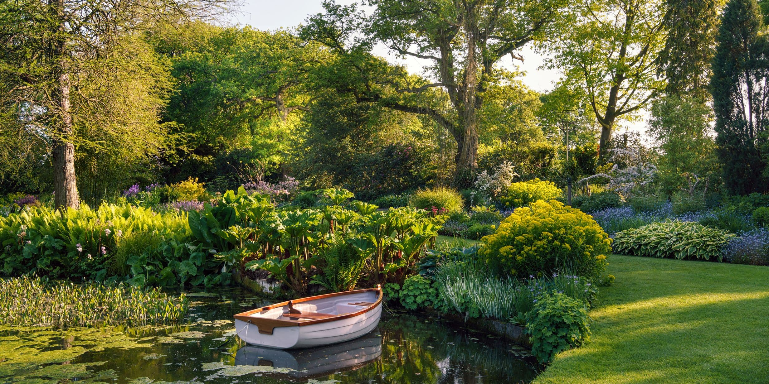 A small wooden boat floats on a tranquil pond surrounded by lush greenery and colorful flowers in a sunlit garden.