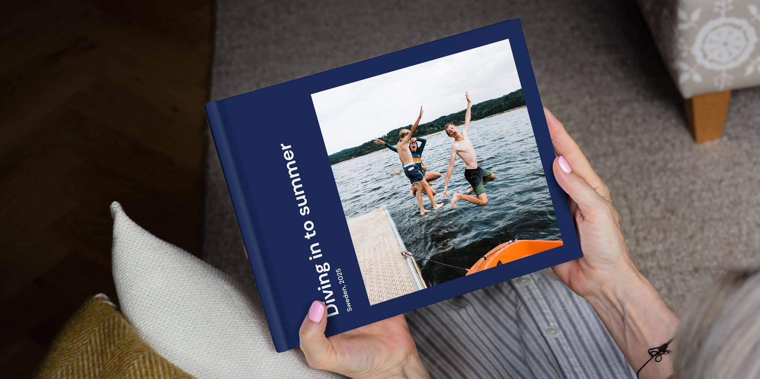 Person holding a photo book titled "Diving in to summer," featuring an image of two people jumping into a lake from a dock.