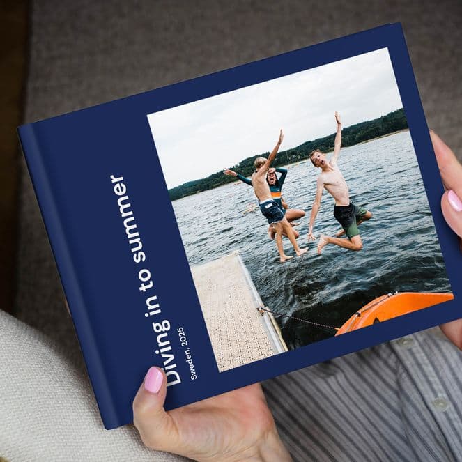 Person holding a photo book titled "Diving in to summer," featuring an image of two people jumping into a lake from a dock.