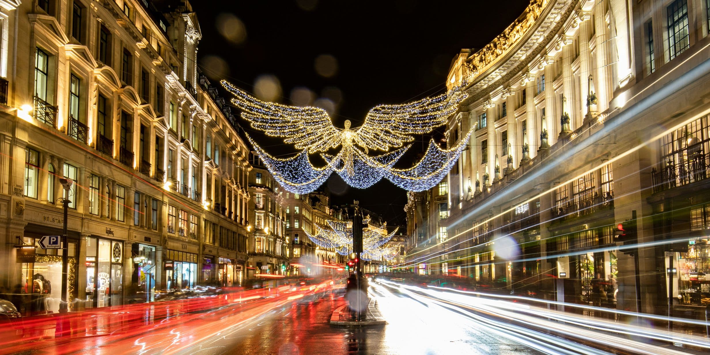 Night scene of a busy city street with angel-shaped Christmas lights overhead, illuminated buildings, and blurred light trails from passing vehicles.
