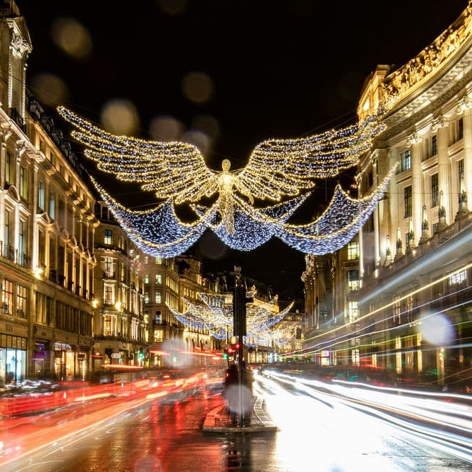 Night scene of a busy city street with angel-shaped Christmas lights overhead, illuminated buildings, and blurred light trails from passing vehicles.
