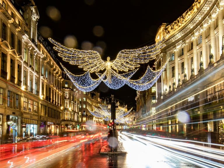 Night scene of a busy city street with angel-shaped Christmas lights overhead, illuminated buildings, and blurred light trails from passing vehicles.