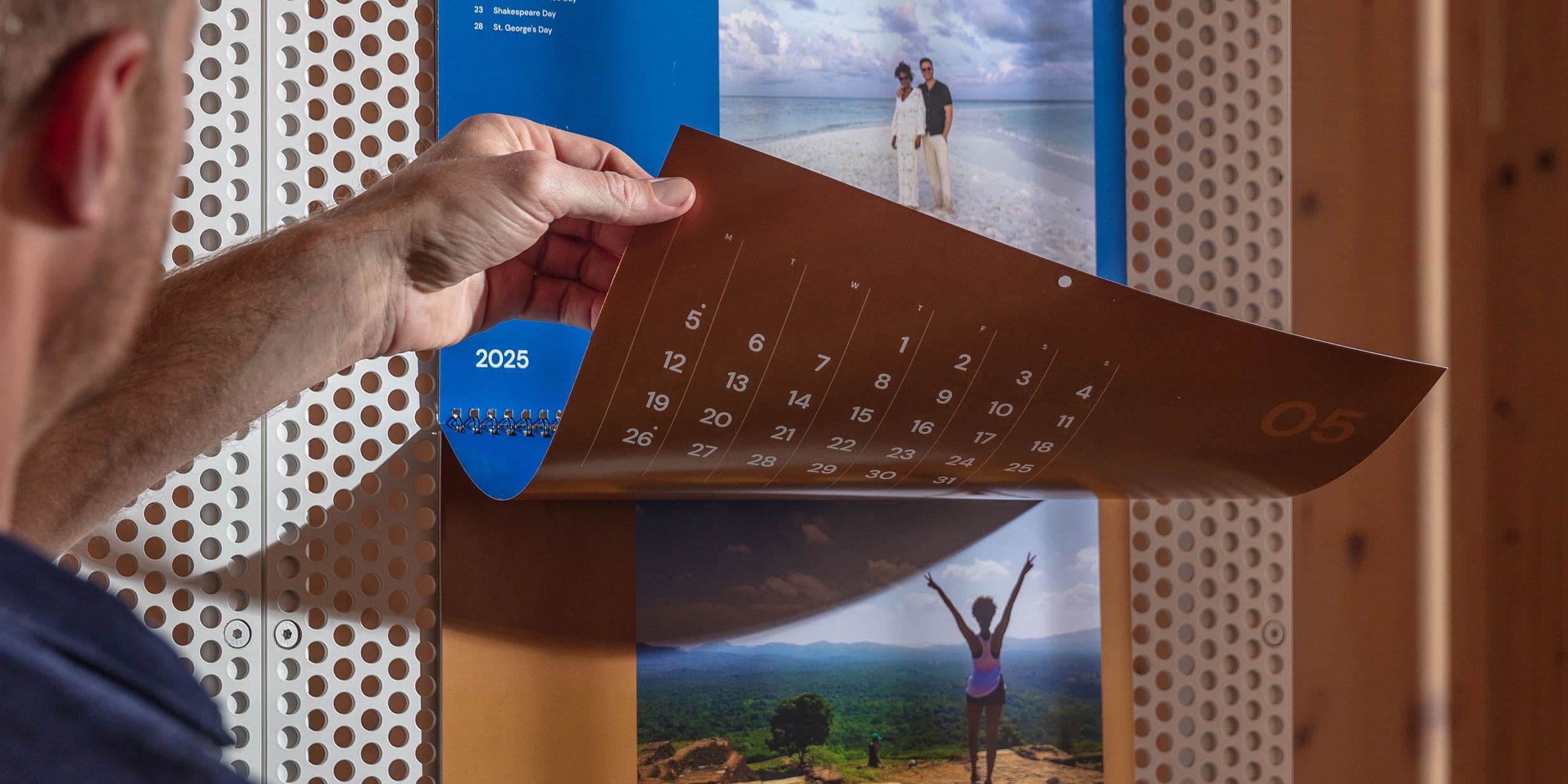 Person flipping calendar page to May 2025, featuring beach and mountain images, on a perforated wall background.