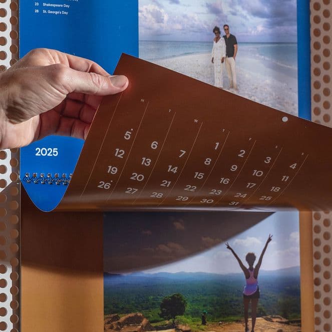Person flipping calendar page to May 2025, featuring beach and mountain images, on a perforated wall background.