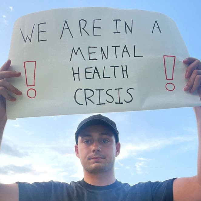 Person holding a sign reading "We are in a mental health crisis" with exclamation marks, against a blue sky background.