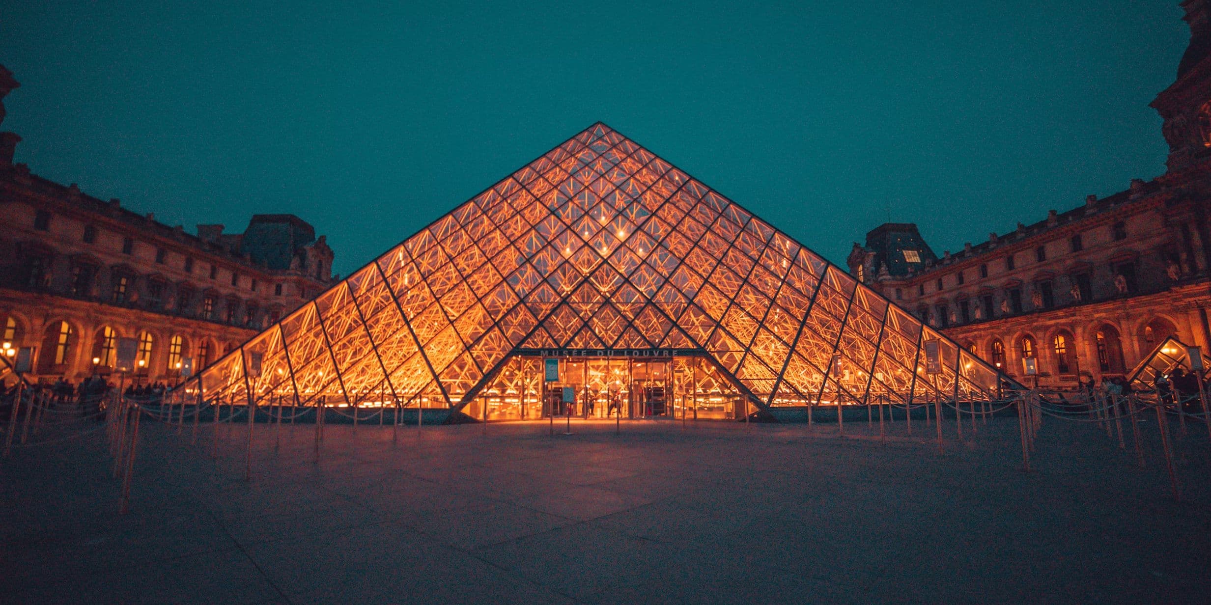 Illuminated glass pyramid entrance of the Louvre Museum at night, surrounded by historic buildings under a dark turquoise sky.