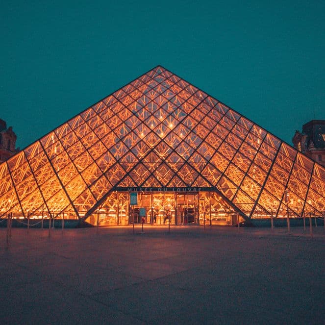 Illuminated glass pyramid entrance of the Louvre Museum at night, surrounded by historic buildings under a dark turquoise sky.