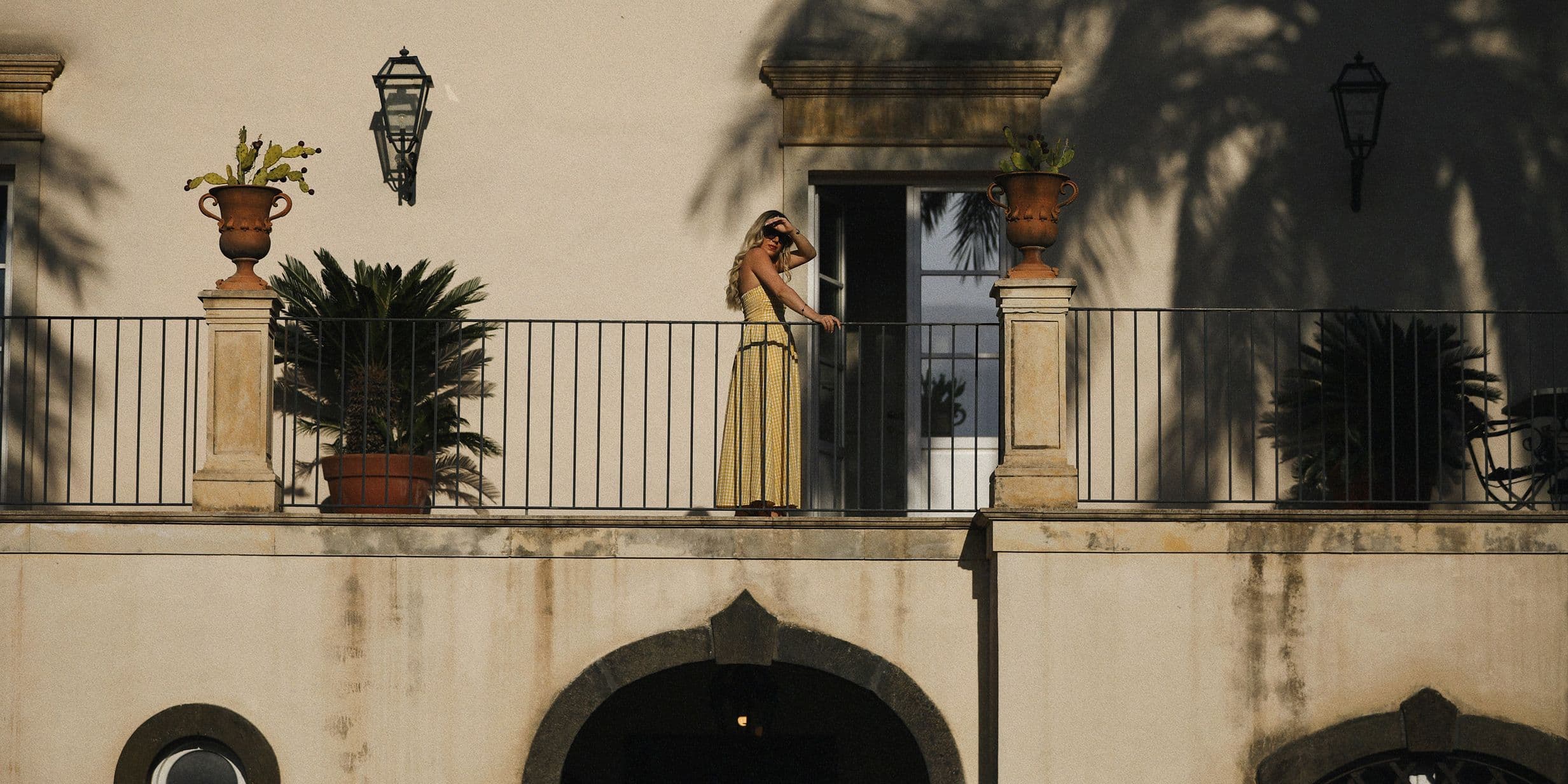 A person in a yellow dress stands on a balcony with potted plants, under the shadow of palm trees, against a beige building.
