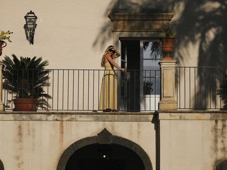 A person in a yellow dress stands on a balcony with potted plants, under the shadow of palm trees, against a beige building.