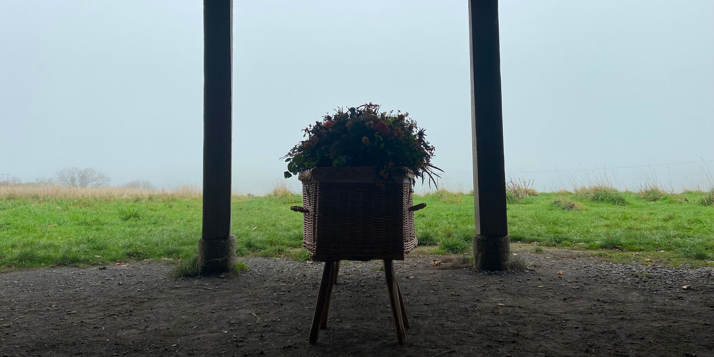 Wicker planter with flowers on a wooden stand centered between two support posts, overlooking a foggy green field.