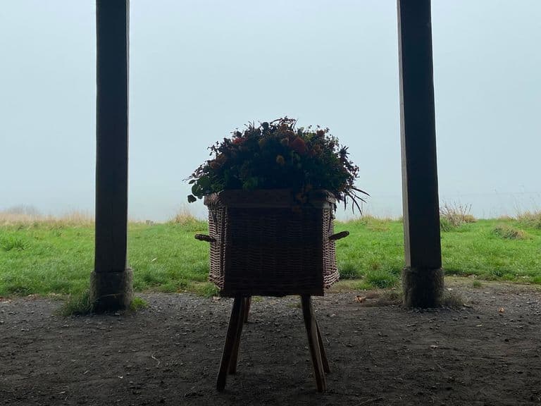 Wicker planter with flowers on a wooden stand centered between two support posts, overlooking a foggy green field.