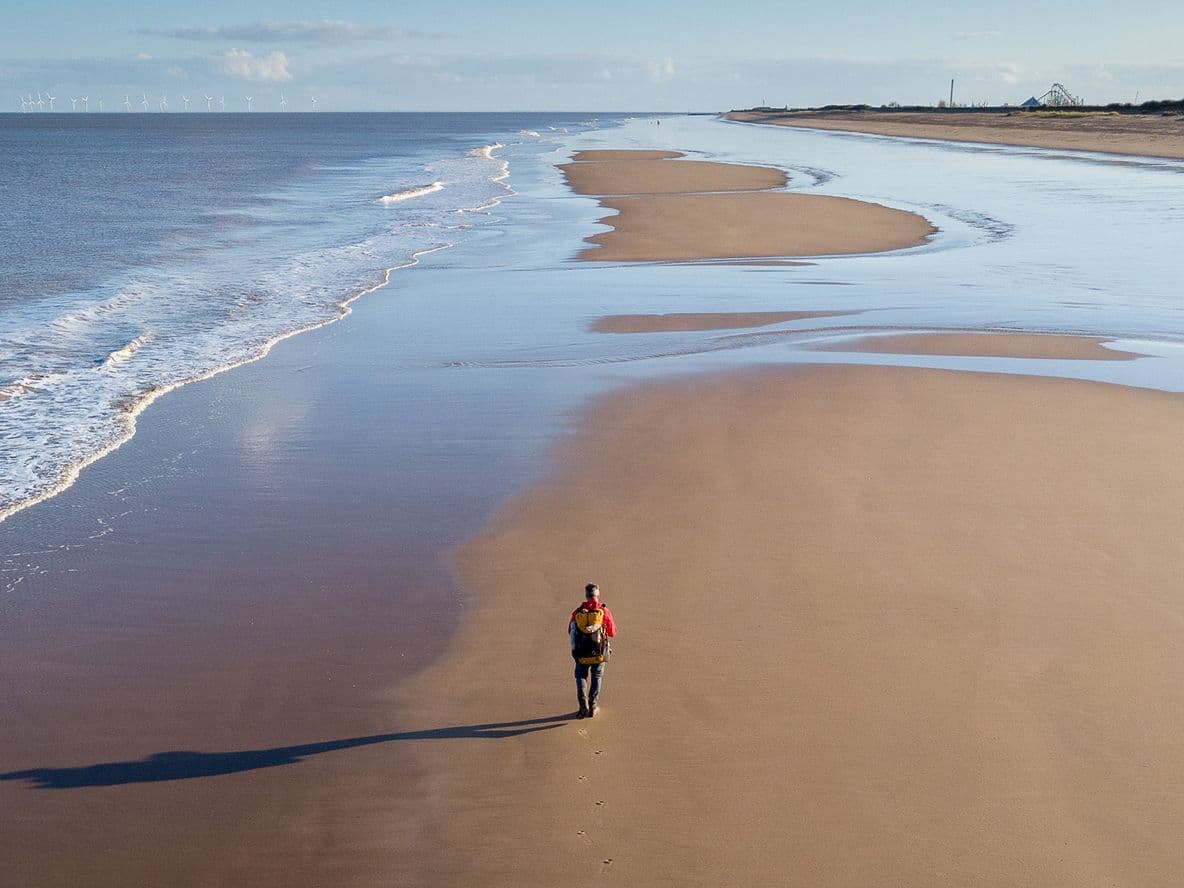 Person walking alone on a vast, empty beach with gentle waves and a clear sky, casting a long shadow on the sand.