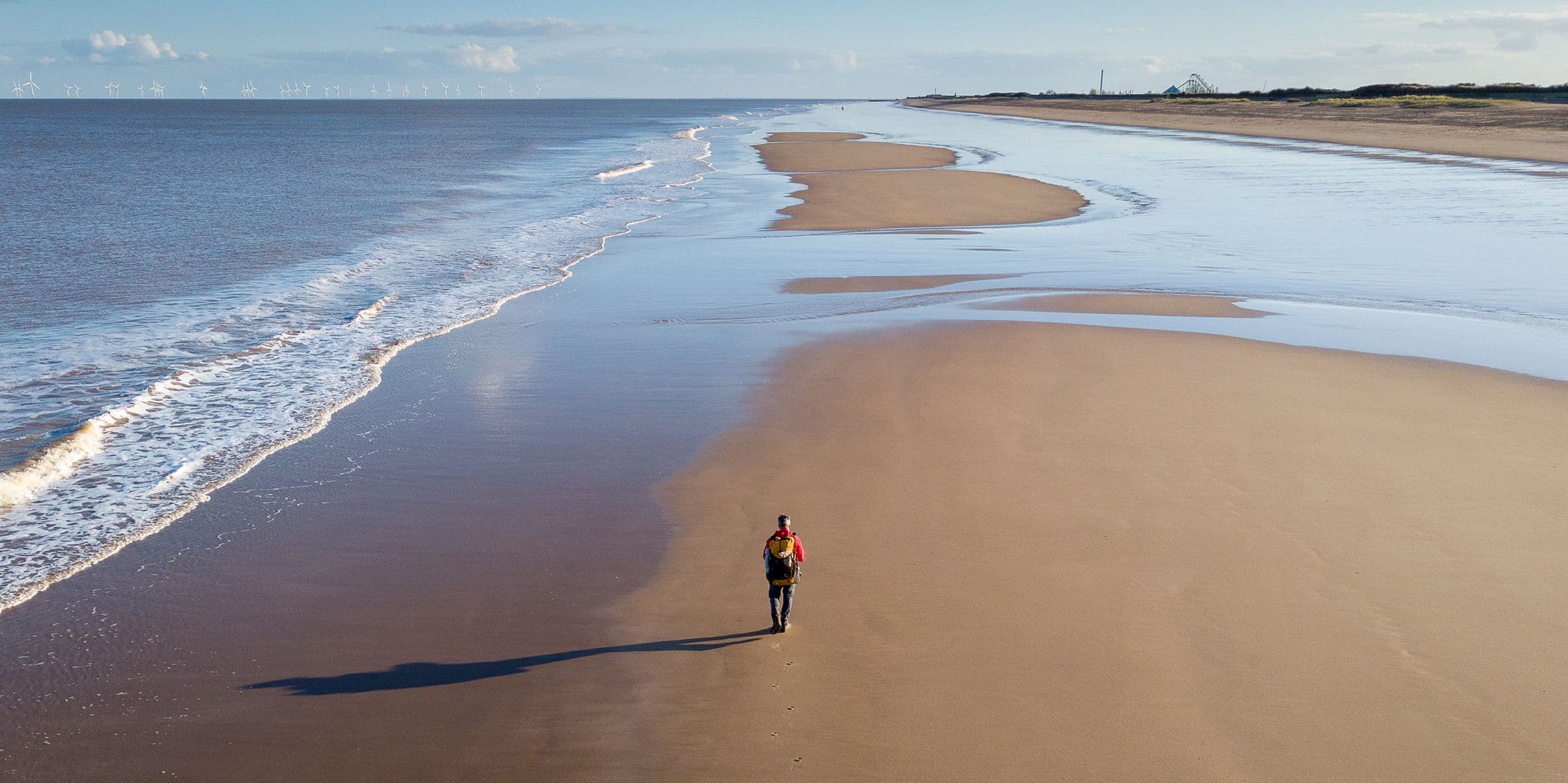 Person walking alone on a vast, empty beach with gentle waves and a clear sky, casting a long shadow on the sand.
