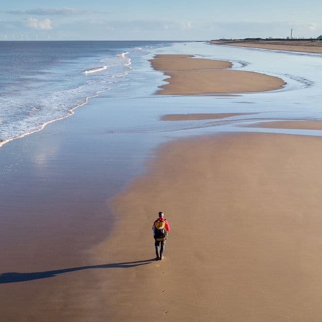 Person walking alone on a vast, empty beach with gentle waves and a clear sky, casting a long shadow on the sand.