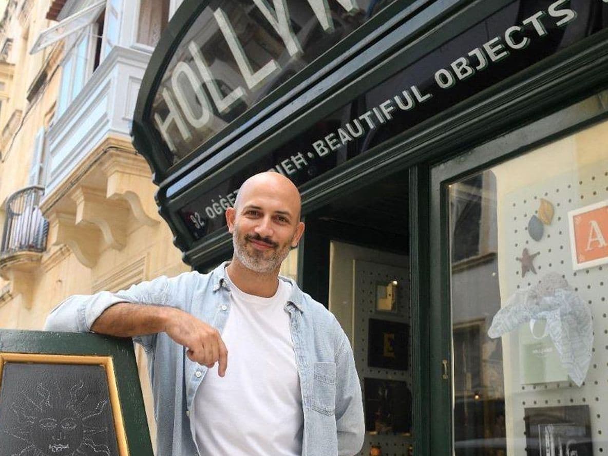 A man stands smiling outside a store named "Hollywood" with a chalkboard sign and display window showing various decorative items.
