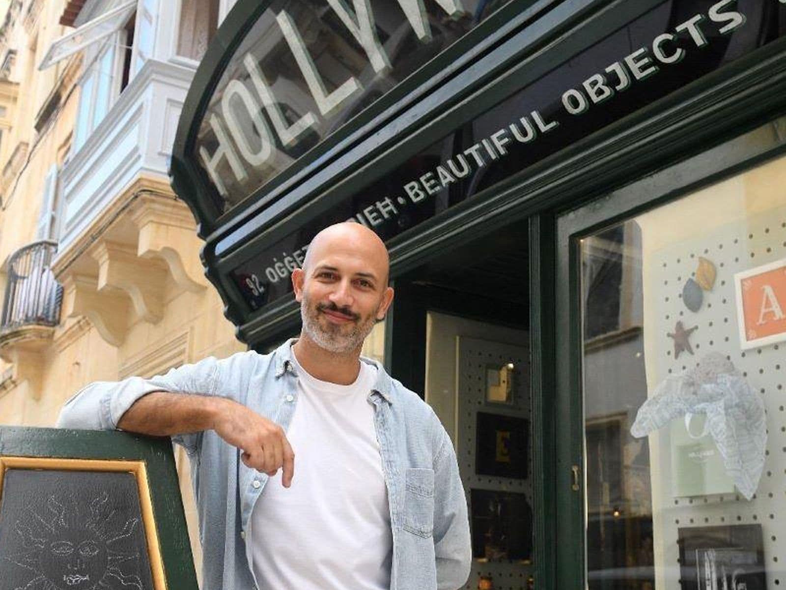 A man stands smiling outside a store named "Hollywood" with a chalkboard sign and display window showing various decorative items.
