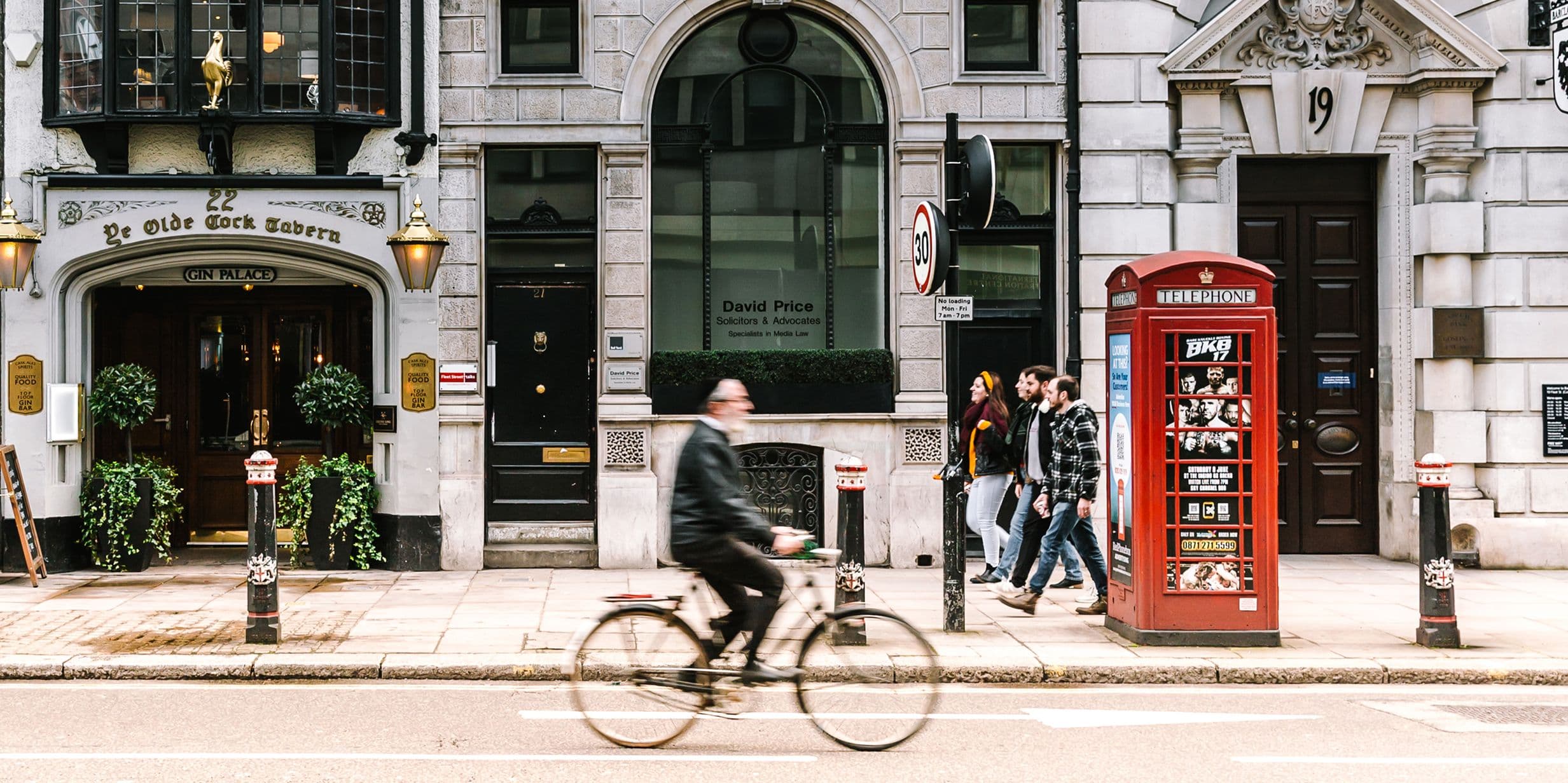 A cyclist passes by a classic red telephone booth and pedestrians on a city street, lined with historic buildings and a pub.