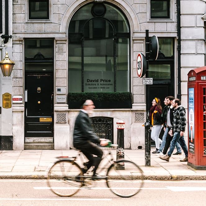A cyclist passes by a classic red telephone booth and pedestrians on a city street, lined with historic buildings and a pub.