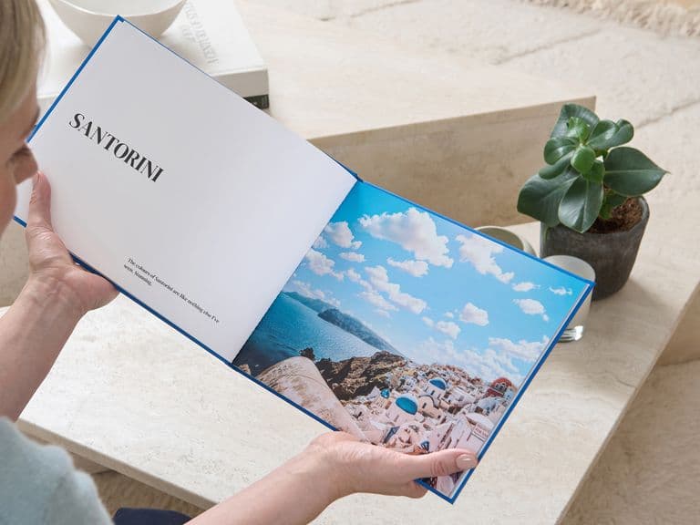 A person holds an open book with a page titled "Santorini" and a scenic photo of a coastline. A small plant is on a table nearby.