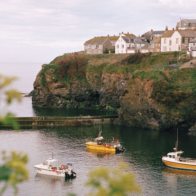 Coastal village with houses on a cliff, overlooking a calm sea with three small boats. Foreground features blurred greenery.
