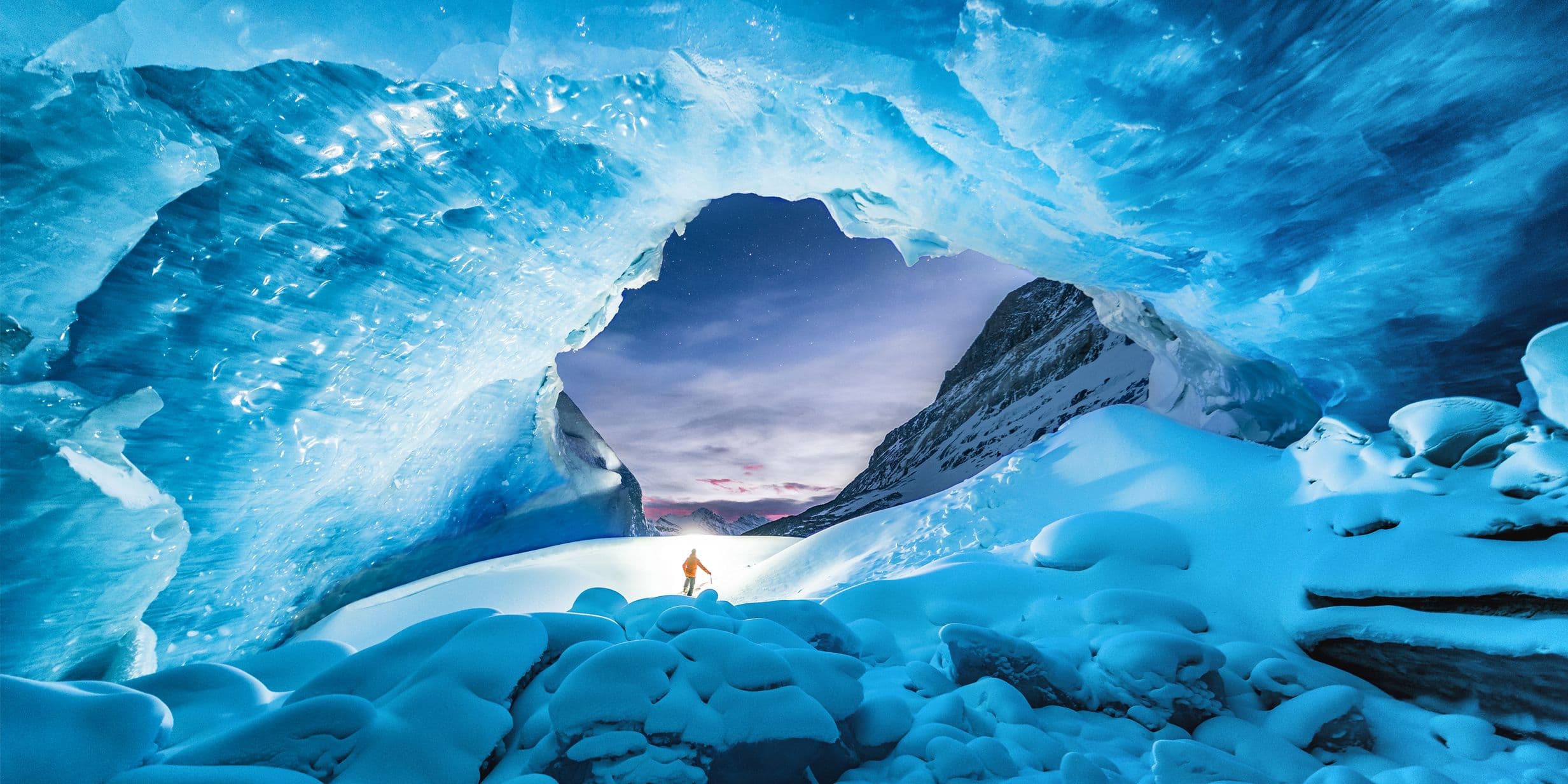 Person in winter clothing stands under a vast, blue ice cave with a view of snowy mountains and a twilight sky in the background.
