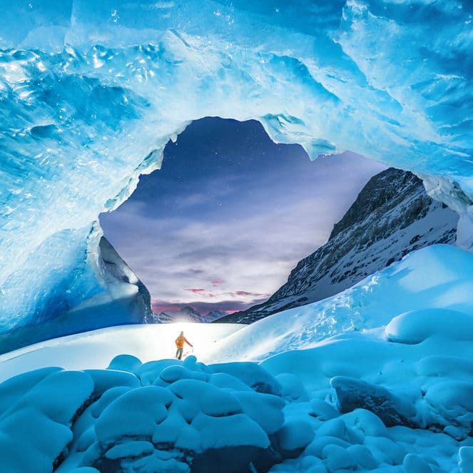 Person in winter clothing stands under a vast, blue ice cave with a view of snowy mountains and a twilight sky in the background.