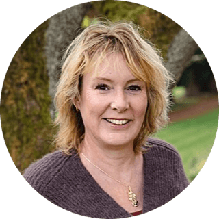 Smiling woman with blonde hair, wearing a brown sweater and necklace, stands outdoors in front of a tree and grass.