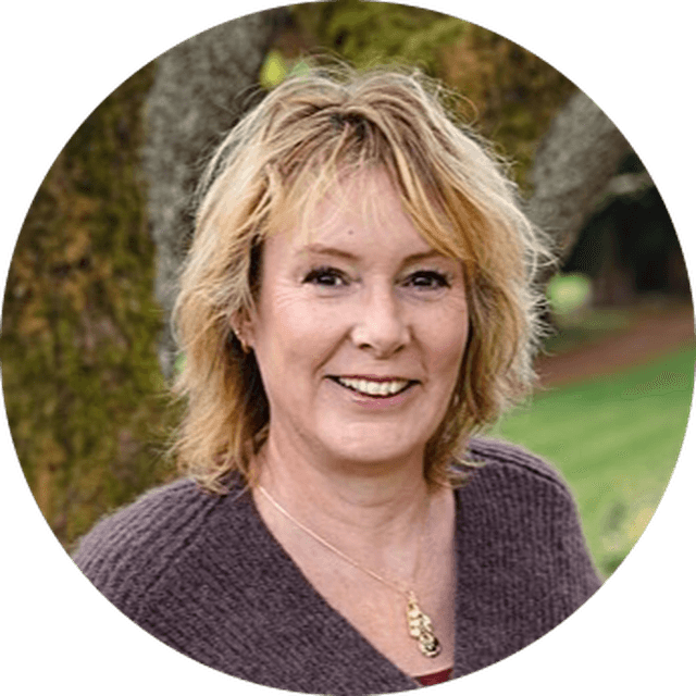 Smiling woman with blonde hair, wearing a brown sweater and necklace, stands outdoors in front of a tree and grass.