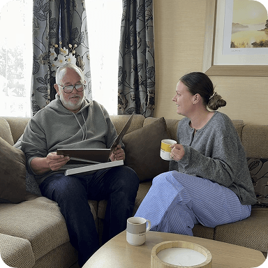Older man and younger woman sitting on a sofa, looking at a photo book. The woman holds a mug, and they appear to be enjoying a conversation.