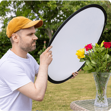 Dan, wearing a cap holds a reflector, adjusting lighting on a vase of colorful roses outdoors.