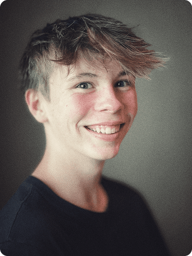Smiling young person with tousled hair, wearing a dark shirt, looking at the camera against a neutral background.